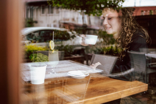 Cute brunette, curly girl sitting in a cafe, drinking coffee and juice. View though the wondow - Photo, Image