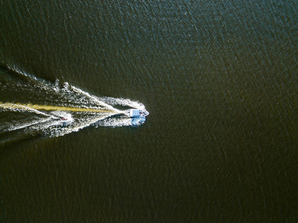 Αεροφωτογραφία τηλεκατευθυνόμενου. Ξυπνήστε σέρφινγκ πίσω από μια βάρκα στο ποτάμι μια ηλιόλουστη καλοκαιρινή μέρα. - Φωτογραφία, εικόνα