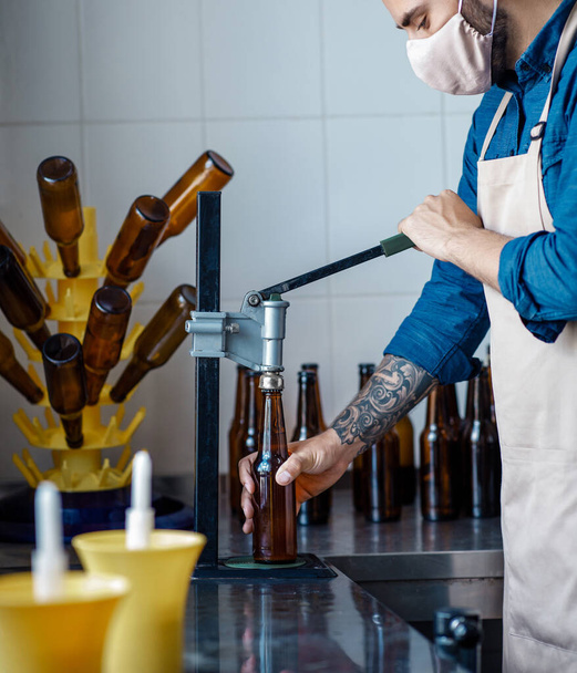 Bira ambalajı, bira fabrikası ve fabrika işi. Önlüklü ve koruyucu maskeli genç çekici işçi şişe tıpası takıyor. - Fotoğraf, Görsel