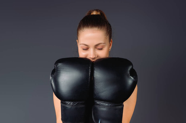 Gözleri kapalı genç bir iş kadını, başarısıyla seviniyor, gri arka planda boks eldivenleri tutuyor. Yüksek kalite fotoğraf - Fotoğraf, Görsel
