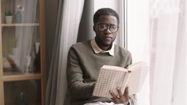 Medium shot van jonge Afrikaanse man het dragen van casual kleding en glazen zitten op vensterbank en lezen boek - Video