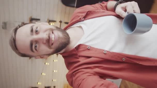 Verticale handheld taille-up shot van vrolijke bebaarde knappe blanke man met uitgestrekte hand wandelen rond keuken thuis en het hebben van vriendelijke prive-gesprek op video oproep - Video