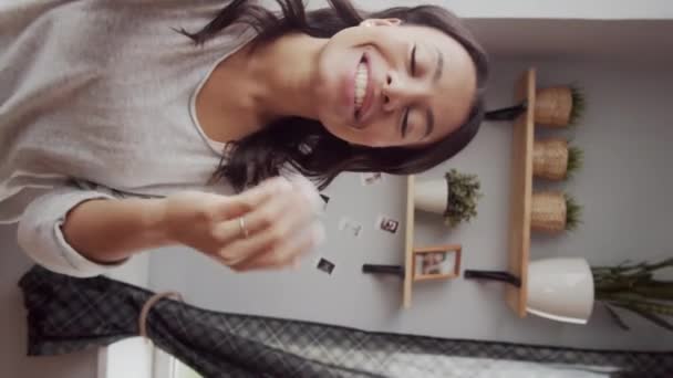 Vertical handheld waist-up shot of vivacious young mixed race woman with genuine toothy smile standing by window at home with outstretched hand and having friendly personal chat on video call - Filmagem, Vídeo