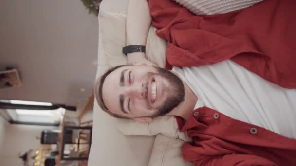 Vertical handheld waist-up shot of vivacious bearded Caucasian man lying on couch at home, holding out outstretched hand and having friendly chat on video call, looking at camera with genuine smile - Felvétel, videó