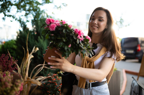 νεαρή όμορφη μελαχρινή γυναίκα απολαμβάνοντας λουλούδια σε γλάστρες στη βεράντα ενός ανθοπωλείου - Φωτογραφία, εικόνα
