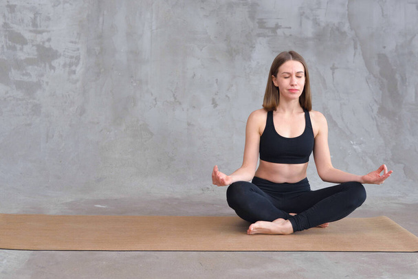 Brązowłosa kobieta w czarnej odzieży sportowej na szarym tle betonu, siedząca w pozycji do jogi i medytacji. Kobieta medytująca w pozycji lotosu. Pojęcie relaksu. Rekreacja i harmonia - Zdjęcie, obraz
