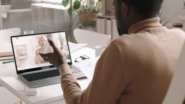 Back view of good-looking African-American businessman sitting at desktop in front of laptop and discussing charts with female business partner by internet connection - Footage, Video