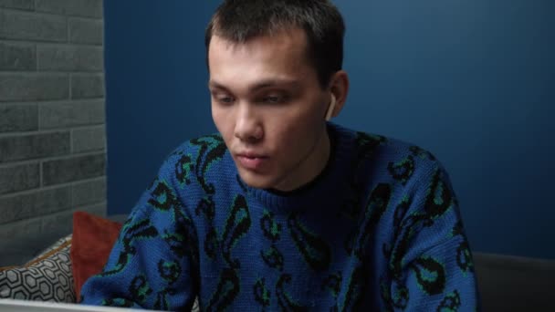 Ein fokussierter, gutaussehender junger Mann aus dem Kaukasus, der Kopfhörer trägt und zu Hause auf dem Sofa seinen Laptop benutzt. Glücklicher Kaukasier, der am Laptop arbeitet, Musik hört und zu Hause auf dem Sofa singt. - Filmmaterial, Video