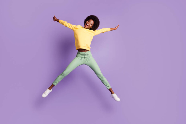 フルボディ写真の屈託のないアフリカ系アメリカ人の女の子ジャンプ身に着けている黄色セーター緑のズボン孤立した上紫色の背景 - 写真・画像