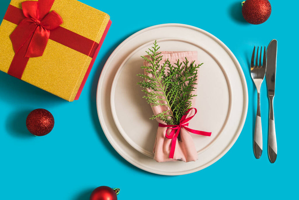 Served table with plate and cutlery for celebration of Christmas and New Year. On plate is napkin with a Christmas tree branch, red balls. Flatlay on blue background with balls, gift box. - Photo, Image