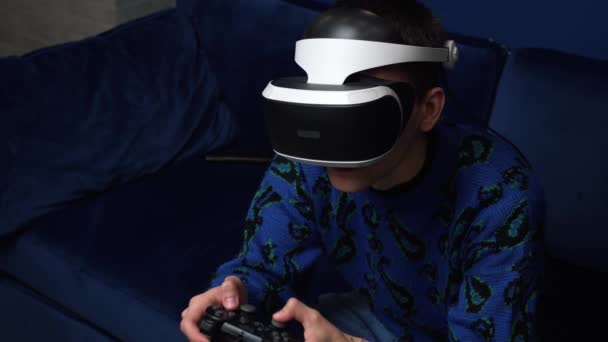 Vonzó izgatott kaukázusi férfi tartja joystick és visel virtuális valóság szemüveg fejhallgató sisak kanapén otthon. Boldogság játékos játék segítségével VR szemüveg otthon. - Felvétel, videó