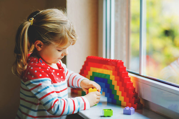 Cute little toddler girl by window create rainbow with colorful plastic blocks during pandemic coronavirus quarantine. Children made and paint rainbows around the world as sign. - Photo, Image
