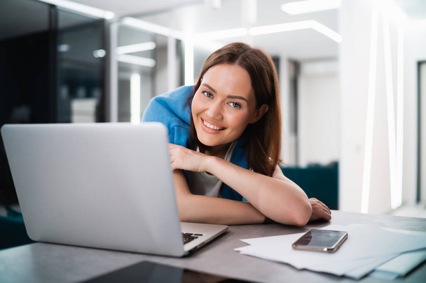 Una joven blogger influencer sonriente que trabaja en casa. Chica feliz trabajando remotamente desde casa sentado en el escritorio con el ordenador portátil y sonriendo a la cámara. - Foto, imagen
