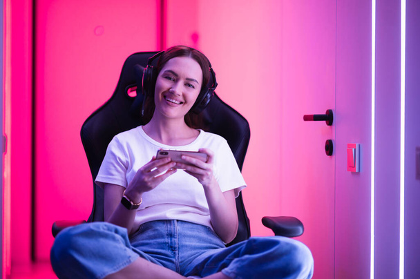 Gamer κορίτσι σε ένα μεγάλο επαγγελματικό ακουστικά μουσικής σε λευκό t-shirt και μπλε τζιν κρατά έξυπνο τηλέφωνο στα χέρια και παίζει το παιχνίδι app στο κινητό τηλέφωνο, χαμογελώντας και κοιτάζοντας κάμερα σε εσωτερικούς χώρους. - Φωτογραφία, εικόνα
