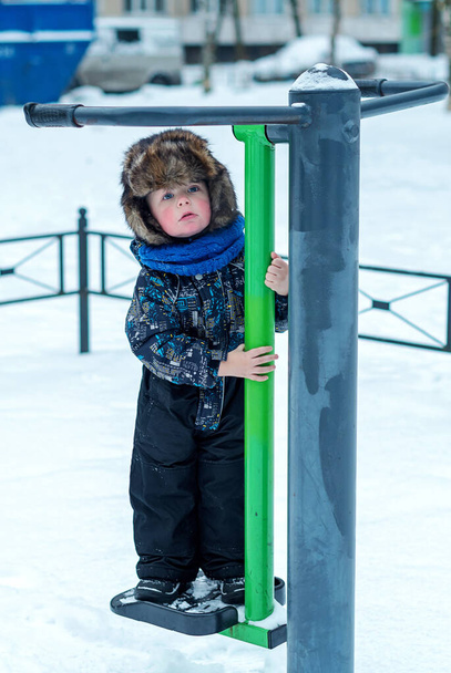3-vuotias poika haalareissa seisoo ulkoiluharjoituskoneessa aikuisille. Talvella kävellä. - Valokuva, kuva