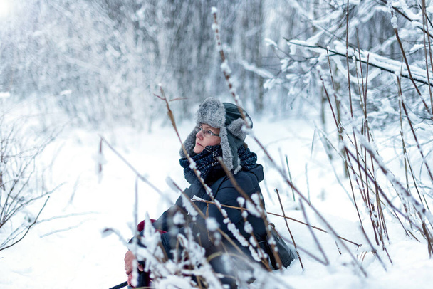 Una mujer se sienta entre los árboles cubiertos de nieve en un bosque de invierno y disfruta de la primera nieve. En un sombrero con orejeras. El concepto de las vacaciones de invierno - Foto, Imagen