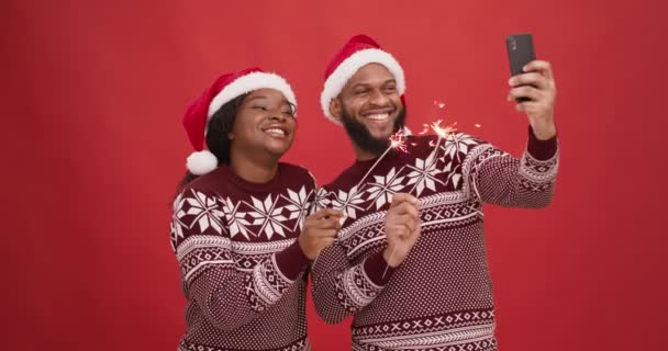 Happy african american couple in santa hats holding fireworks and recording video on smartphone, red studio background - Footage, Video