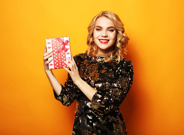 Portrait of a happy smiling woman in dress holding present box isolated over yellow background - Photo, image