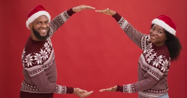 Cooles Weihnachtsangebot. Glückliches afrikanisch-amerikanisches Paar in Weihnachtsmützen, die Rahmen mit Händen, rotem Hintergrund, leerem Raum herstellen - Filmmaterial, Video