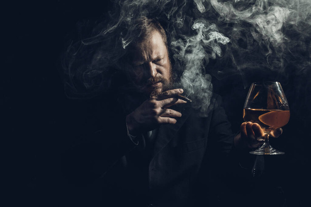 Solid confident bearded man in suit with glass of whisky and cigar with fume - Foto, afbeelding