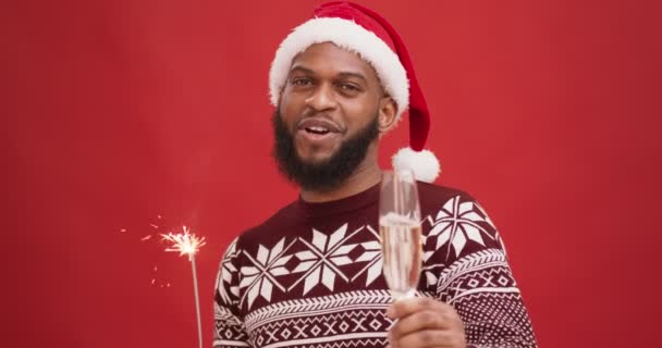 Giovane uomo nero in cappello di Babbo Natale in possesso di bicchiere di champagne e luce del bengala, dicendo Buon Natale e Felice Anno Nuovo - Filmati, video