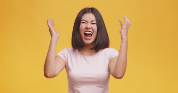 Молодая яростная азиатка кричит громко и пожимает руки, чувствует ярость и гнев, оранжевый фон студии - Кадры, видео