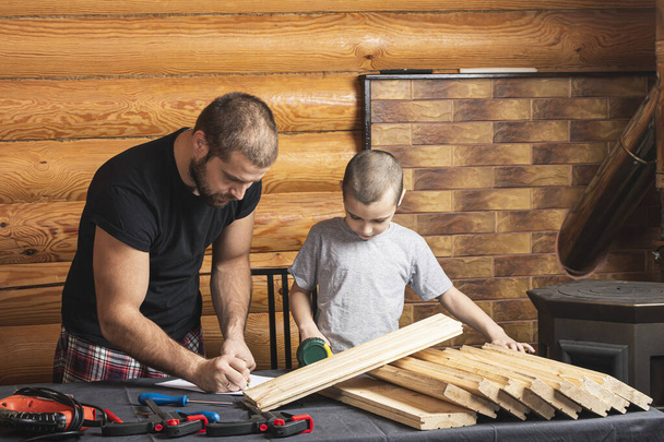 Dad and son are drawing on paper, planning how to build a bird house, tools and a beam on the table in the workshop. Carpentry training concept for kids - Photo, Image