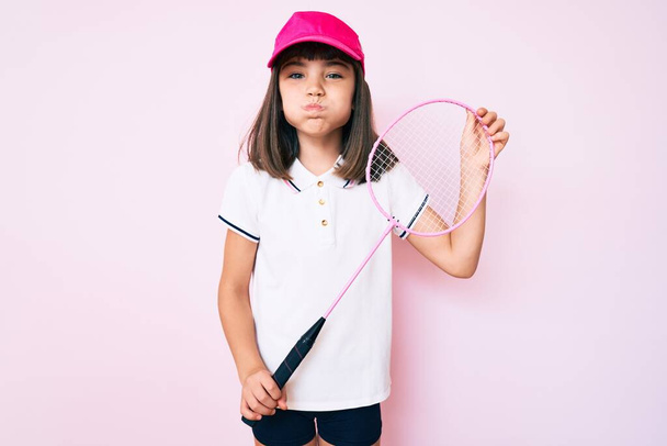 Młoda dziewczynka z hukiem trzymająca rakietę badmintona nadyma policzki z zabawną twarzą. usta napompowane powietrzem, łapie powietrze.  - Zdjęcie, obraz