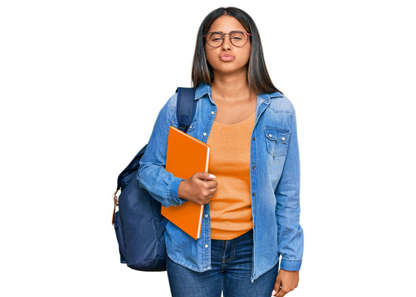 Young latin girl wearing student backpack and holding books looking at the camera blowing a kiss on air being lovely and sexy. love expression.  - Foto, Bild