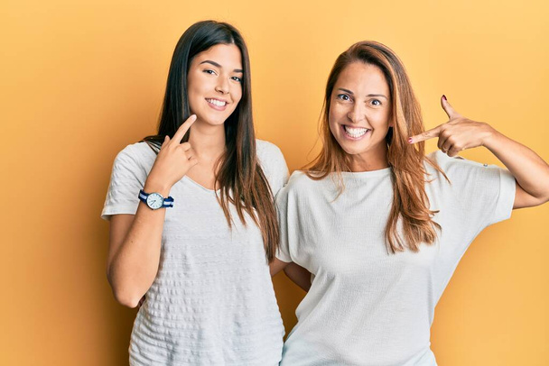 Spaanse familie van moeder en dochter dragen casual witte tshirt lachende vrolijke tonen en wijzen met vingers tanden en mond. tandheelkundige gezondheidszorg.  - Foto, afbeelding