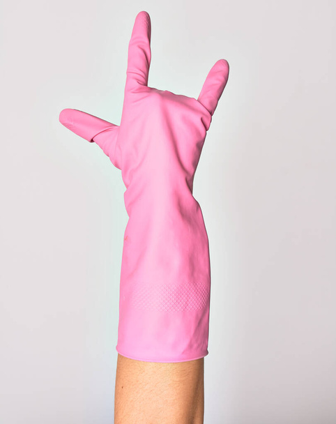 Ręka młodej białej kobiety noszącej różową rękawicę czyszczącą pokazującą palec ze znakiem rogów. Robiąc symbol rocka na pojedynczym białym tle - Zdjęcie, obraz