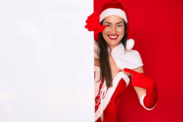 Молодая женщина в костюме Санта-Клауса держит пустой баннер указывая пальцем на одного улыбающегося счастливого и гордого  - Фото, изображение
