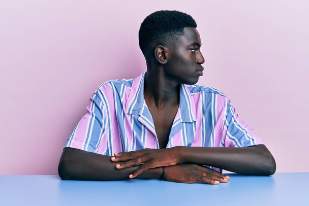 Jeune homme afro-américain portant des vêtements décontractés assis sur la table regardant vers le côté, pose de profil relax avec le visage naturel avec sourire confiant.  - Photo, image