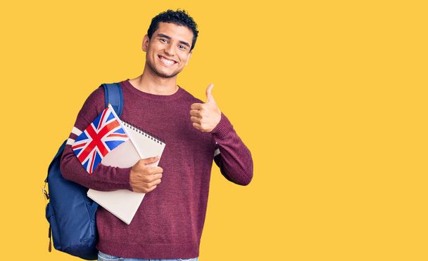 Hispano guapo joven intercambio estudiante sosteniendo Reino Unido bandera sonriendo feliz y positivo, pulgar hacia arriba haciendo excelente y aprobación signo  - Foto, imagen