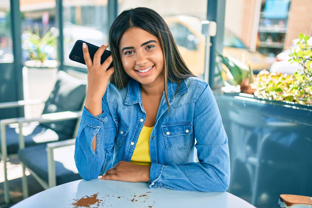 Młoda Latynoska uśmiechnięta szczęśliwa słuchając wiadomości audio za pomocą smartfona na tarasie kawiarni. - Zdjęcie, obraz