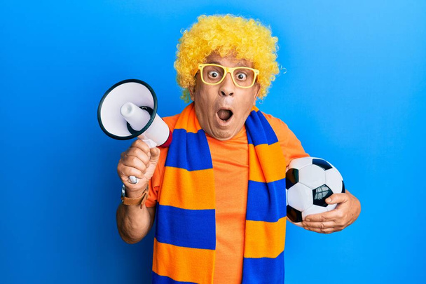 Hombre hispano mayor hooligan fútbol animando juego celebración de pelota y megáfono miedo y conmocionado con sorpresa y expresión asombrada, miedo y cara emocionada.  - Foto, Imagen