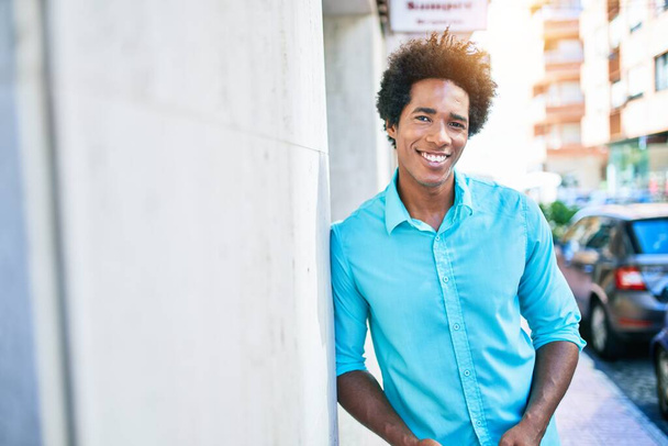 Молодой красивый африканский американец в повседневной одежде улыбается счастливо. Прислонившись к стене с улыбкой на лице на городской улице. - Фото, изображение