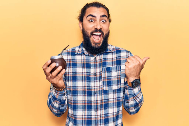 Junger arabischer Mann trinkt Mate-Infusion und zeigt mit dem Daumen zur Seite und lächelt glücklich mit offenem Mund  - Foto, Bild