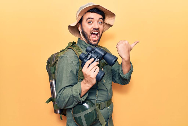 Jovem hispânico vestindo roupas de explorador segurando binóculos apontando o polegar para o lado sorrindo feliz com a boca aberta  - Foto, Imagem