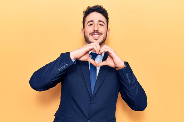 Joven hombre hispano vistiendo traje sonriendo en amor haciendo forma de símbolo del corazón con las manos. concepto romántico.  - Foto, Imagen