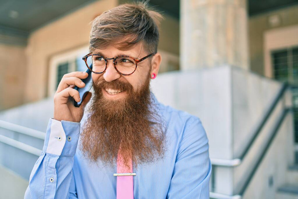 若い赤毛のビジネスマンが笑顔で市内に立っているスマートフォンで話しています. - 写真・画像