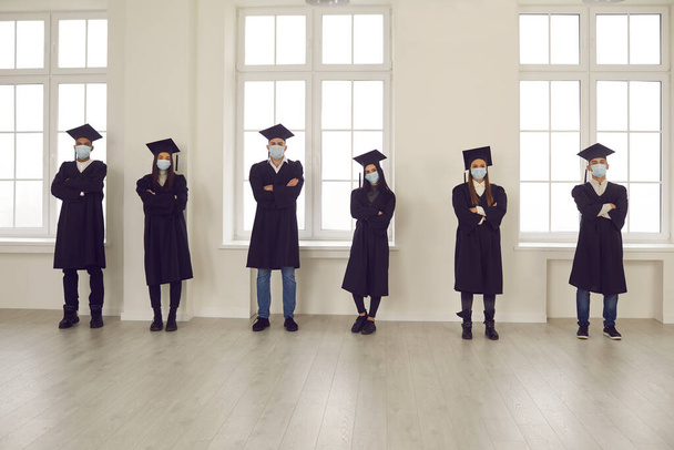 Students in graduation robes and face masks standing in university room, keeping social distance - Fotoğraf, Görsel