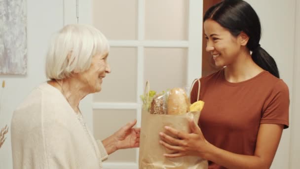 Junge asiatische Pflegerin bringt Tüte mit Lebensmitteln zur Rentnerin nach Hause. Glückliche Seniorinnen umarmen sich und sagen Danke - Filmmaterial, Video