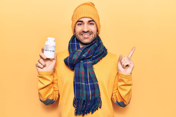 Young handsome bald man wearing winter clothes holding jar of pills smiling happy pointing with hand and finger to the side  - Foto, imagen