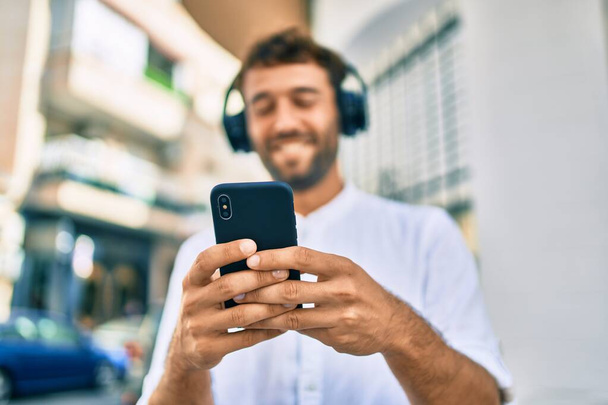 Красивий чоловік з бородою в повсякденній білій сорочці на сонячний день посміхається щасливий на відкритому повітрі, використовуючи смартфон, що слухає музику в навушниках
 - Фото, зображення