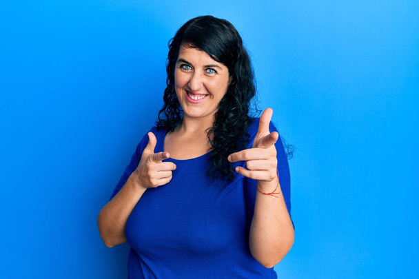プラスサイズのブルネットの女性は、幸せと面白い顔を持つカメラに指を指してカジュアルな青いシャツを着ています。良いエネルギーと振動は.  - 写真・画像