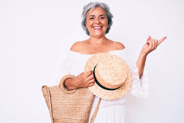 Senior hispanic grey- haired woman wearing casual clothes holding summer hat and handbag smiling happy pointing with hand and finger to the side  - Fotoğraf, Görsel