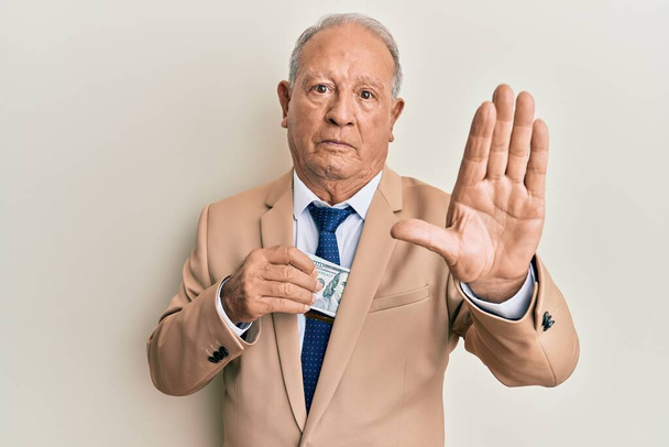 Hombre caucásico mayor ahorrando dinero soborno con la mano abierta haciendo stop sign con expresión seria y segura, gesto de defensa  - Foto, imagen