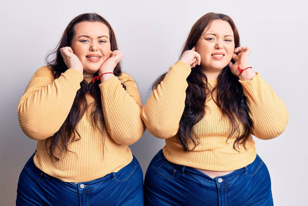 Young plus size twins wearing casual clothes covering ears with fingers with annoyed expression for the noise of loud music. deaf concept.  - Photo, Image