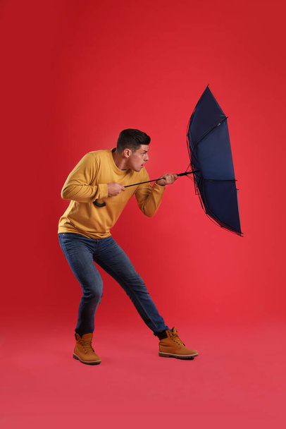 Emotional man with umbrella caught in gust of wind on red background - Photo, Image